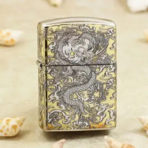 Gold-toned metal lighter with intricate black dragon design across front and sides, standing on a sandy surface with seashells nearby.