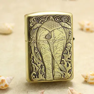 Decorative brass lighter with a raised elephant design, standing on sand with seashells nearby (beach motif).