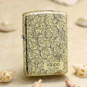 Zippo lighter in gold with ornate scrolling floral pattern, standing on sand with seashells nearby.