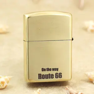 KZ160-01 Brass Zippo-style lighter standing upright with 'On the way Route 66' printed on its front, on a sandy surface with tiny seashells nearby.