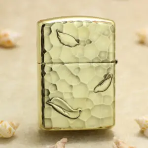 KZ204-01 Gold Zippo-style lighter with a hammered textured finish and leaf motifs on a sandy background with seashells nearby.