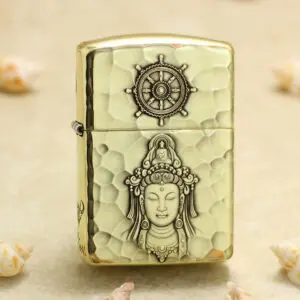 Brass hammered-light metal lighter with a ship-wheel emblem on top and a carved Buddha figure on the body, lying on sand with shells nearby.