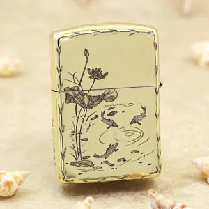 Gold Zippo-style lighter with a koi pond and lotus flower design on a pale background, set on sand with seashells nearby.