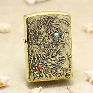 Gold-tone Zippo lighter with an intricate dragon engraving, featuring a turquoise eye and red orb, on a sandy surface with seashells nearby.