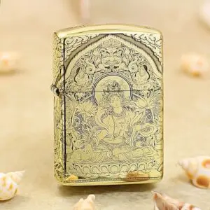 Gold Zippo-style lighter with an intricate Buddhist deity engraving and floral patterns on a sandy surface.