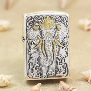 Metal Zippo lighter with an embossed elephant design in gray and gold accents, standing among seashells on a sandy surface.