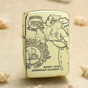 Gold Zippo lighter with vintage illustration and the text 'Since 1932' and 'American Classic' displayed on the front, resting on a sandy surface.