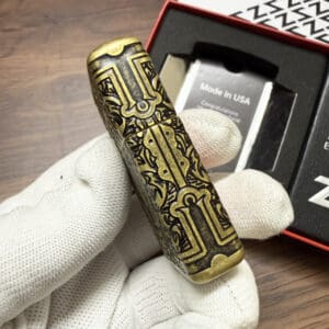 Ornate brass cylinder with intricate black engraving held by a gloved hand, packaging in the background reading 'Made in USA'.