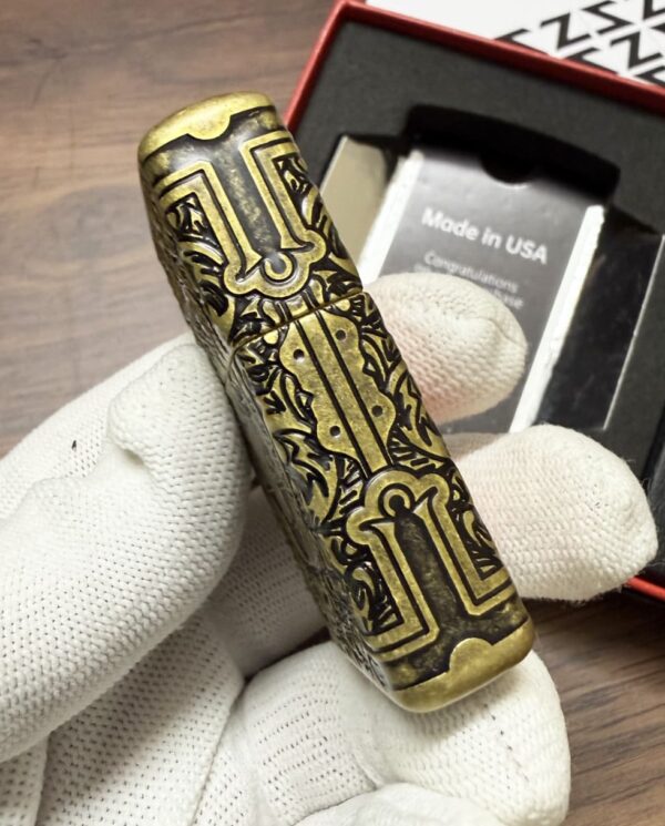 Ornate brass cylinder with intricate black engraving held by a gloved hand, packaging in the background reading 'Made in USA'.