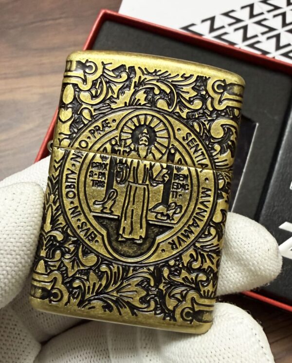 Brass ornate lighter with a circular religious medallion and floral engravings, held by a gloved hand.