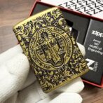 Brass Zippo lighter with ornate old-world engravings and a central circular emblem held by gloved hand on a wooden surface.