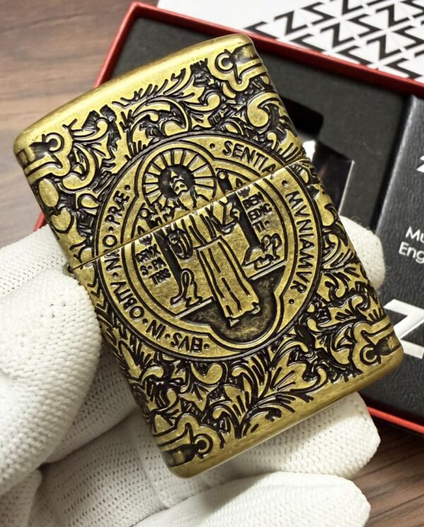 Brass Zippo lighter with ornate old-world engravings and a central circular emblem held by gloved hand on a wooden surface.
