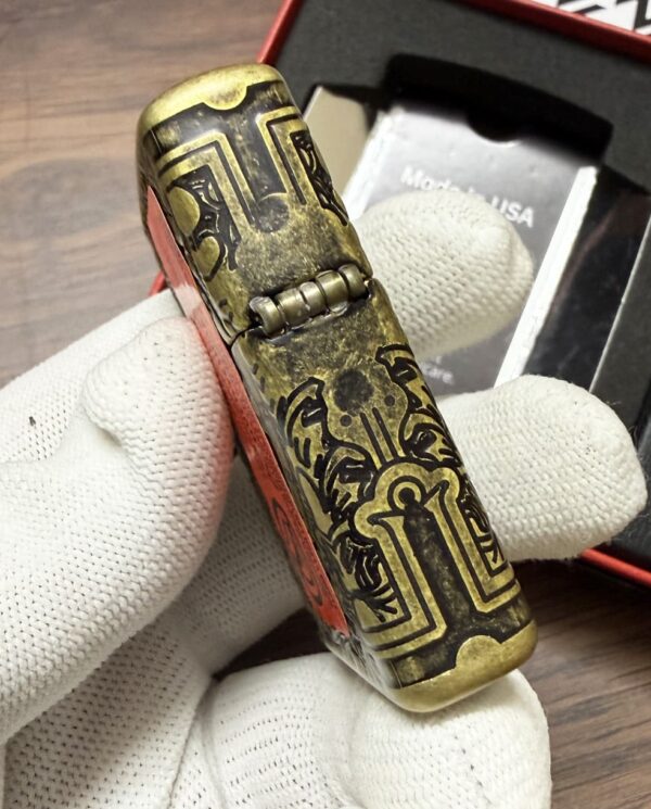 Ornate brass lighter with black engravings held by a gloved hand, the hinge visible.