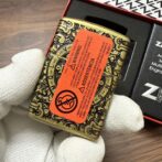 Ornate brass tin being held by gloved hand, orange warning label covering its patterned surface as part of a packaging box nearby.