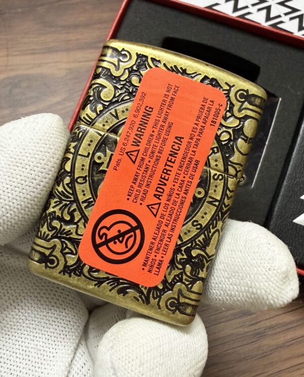 Ornate brass tin being held by gloved hand, orange warning label covering its patterned surface as part of a packaging box nearby.