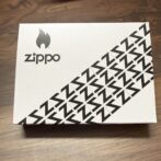 White Zippo box on a wooden surface, featuring the Zippo flame logo and a diagonal pattern of black Z shapes along the bottom-right edge.