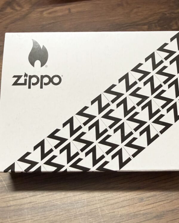 White Zippo box on a wooden surface, featuring the Zippo flame logo and a diagonal pattern of black Z shapes along the bottom-right edge.