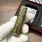Brass engraved cylindrical lighter held by gloved hand, box labeled Made in USA in background.