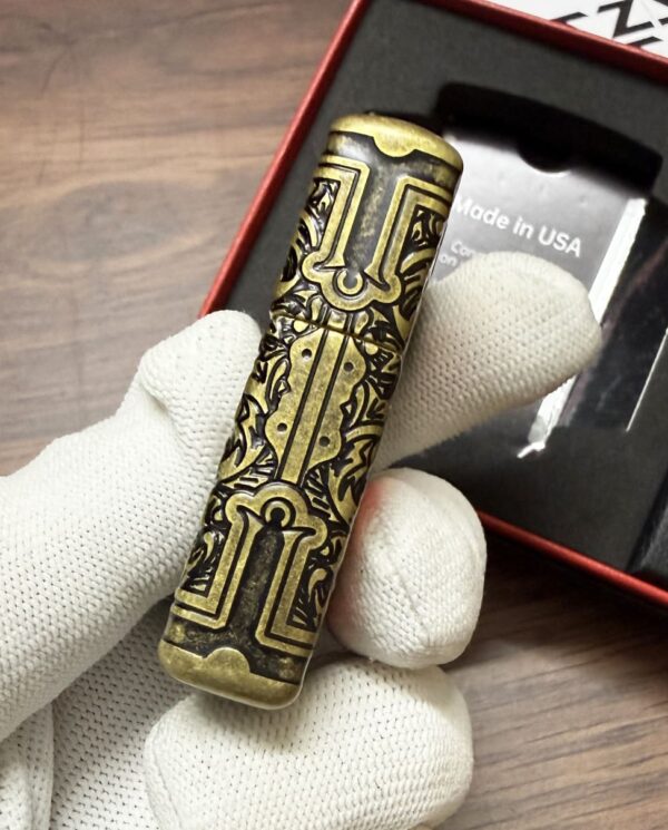 Brass engraved cylindrical lighter held by gloved hand, box labeled Made in USA in background.