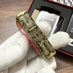 Close-up of an ornate brass cylindrical object with engraved geometric designs, held by gloved hands; a packaging box is visible in the background.