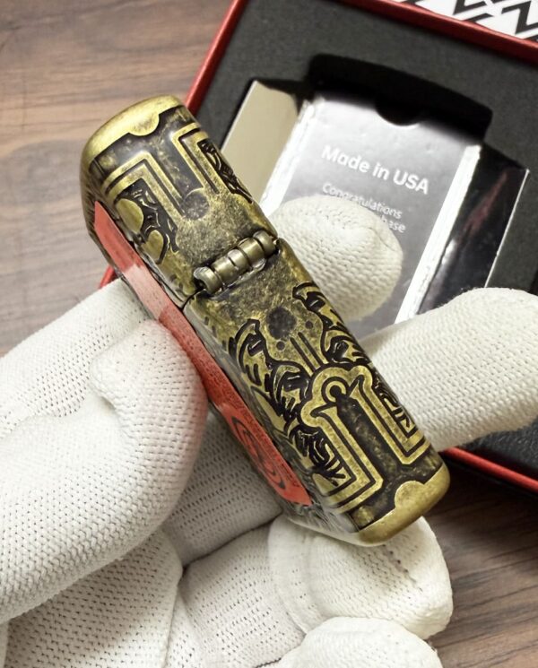 Close-up of an ornate brass cylindrical object with engraved geometric designs, held by gloved hands; a packaging box is visible in the background.