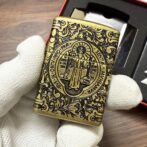 Ornate brass Zippo lighter with a circular saint engraving and floral scrollwork, held by gloved hands (box in background).