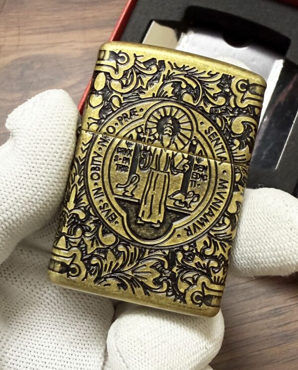 Ornate brass Zippo lighter with a circular saint engraving and floral scrollwork, held by gloved hands (box in background).