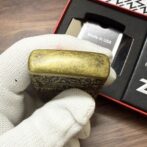Engraved brass lighter being held by a gloved hand, with 'Made in USA' packaging visible in the background.
