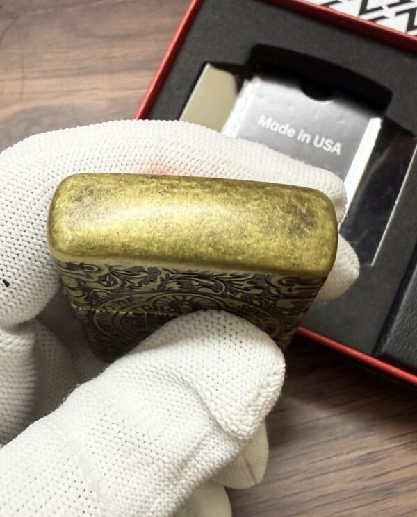 Engraved brass lighter being held by a gloved hand, with 'Made in USA' packaging visible in the background.