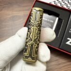 Ornate brass cylindrical object with intricate black engravings held by a gloved hand, packaging visible in the background.