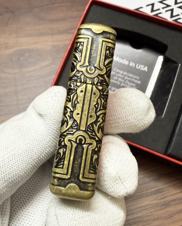 Ornate brass cylindrical object with intricate black engravings held by a gloved hand, packaging visible in the background.