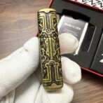 Ornate brass lighter with intricate black engraving, held by a gloved hand with a box visible in the background.
