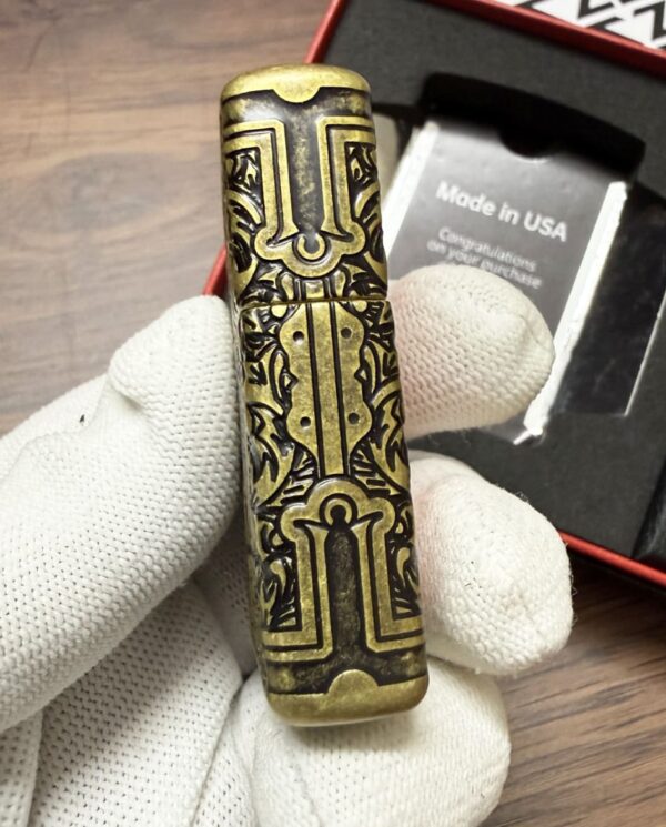 Ornate brass lighter with intricate black engraving, held by a gloved hand with a box visible in the background.