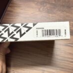 White cardboard box with black geometric pattern, held by hand, showing a barcode sticker on the side.