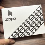 White Zippo box with black Zippo logo and flame icon; diagonal geometric pattern on the lower right corner.