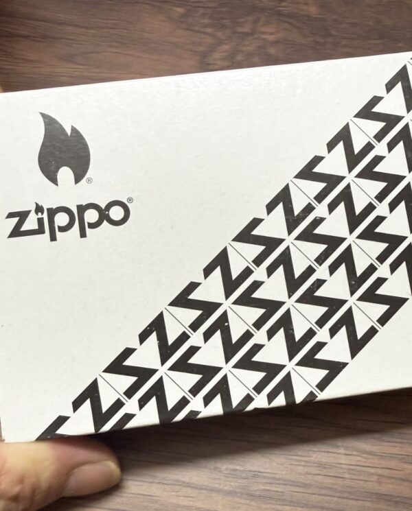 White Zippo box with black Zippo logo and flame icon; diagonal geometric pattern on the lower right corner.