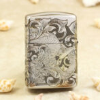 Silver lighter with an intricate dragon and swirling floral engravings on the front, shell-colored background nearby.