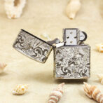 Ornate engraved lighter on sandy beach with seashells nearby, lid open.