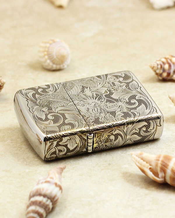 KZ143-04 Metallic cigarette case with ornate floral engraving on a sandy surface, surrounded by seashells.