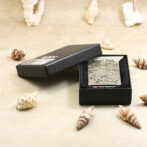 Open black box showing a silver engraved lighter on a foam insert, surrounded by seashells on sand.