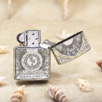 Ornate metal lighter with detailed engravings, open lid, resting on a sandy surface with seashells nearby.