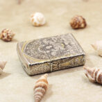 Ornate silver lighter with detailed floral engravings, surrounded by seashells on sandy surface.