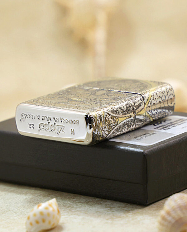 KZ144-05 Engraved metal Zippo lighter with intricate pattern resting on a black box, surrounded by small seashells on a sandy surface.
