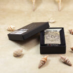 Engraved Zippo-style lighter case in a black presentation box on a sandy surface with seashells nearby.