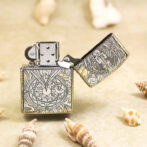 Metal lighter with intricate engraved designs (geometric and dragon motifs) on a sandy surface, lid open.