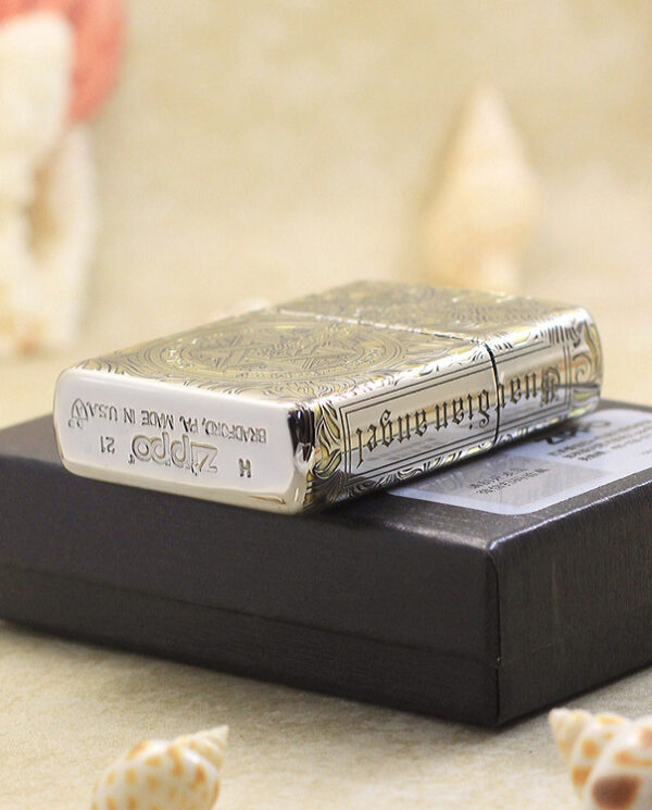 KZ145-05 Shiny silver Zippo lighter with ornate engravings resting on a dark black box on a sandy surface with blurred shells in the background.