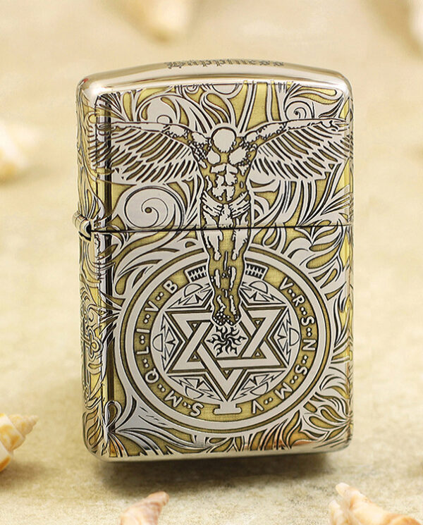 KZ145 Metal lighter with an ornate engraving: a winged figure above a circular geometric emblem in gold and silver tones on a sandy background gaded with seashells.