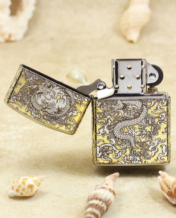 KZ146-02 Gold-toned Zippo-style lighter with an intricate dragon engraving, lid open, set on sandy beach with seashells nearby.