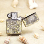 Open, dragon-engraved metal lighter resting on sand among seashells.