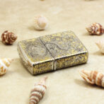 Ornate gold pocket lighter with intricate engravings on a sandy surface among seashells